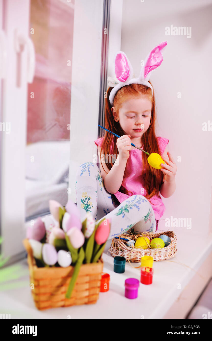 Una piccola bella ragazza con i capelli rossi con orecchie di coniglio sul suo capo è la pittura delle uova per un cesto di Pasqua sul davanzale contro la finestra Foto Stock