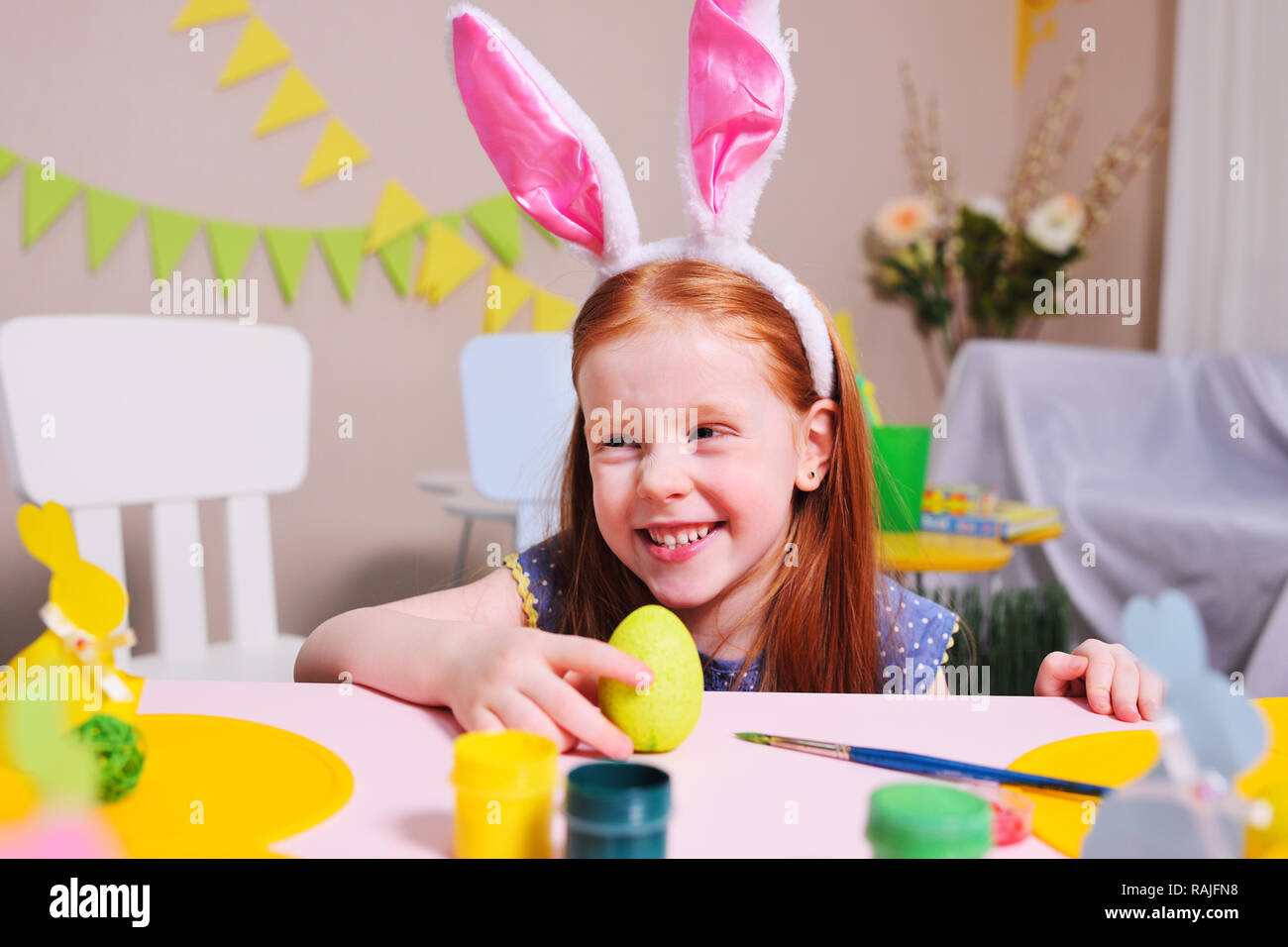 Allegro bambino attivo la ragazza con i capelli rossi e orecchie di coniglio dipinge un uovo con vernice gialla su uno sfondo di pasqua decor e sorrisi. Foto Stock