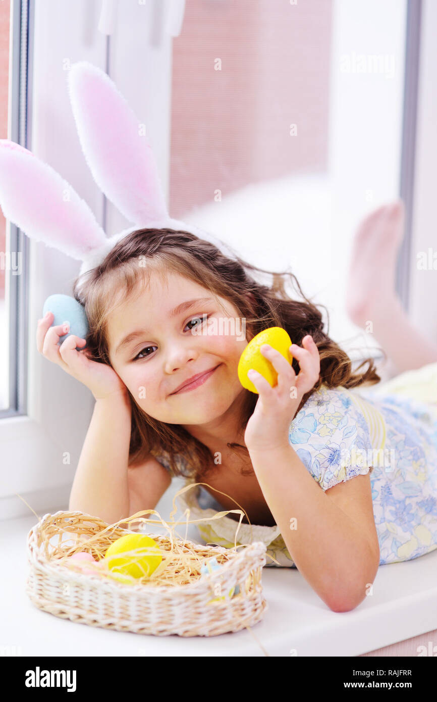 Ragazza con orecchie di coniglio con uova dipinte in mani sorridente sullo sfondo del cesto di pasqua seduto vicino a finestra Foto Stock