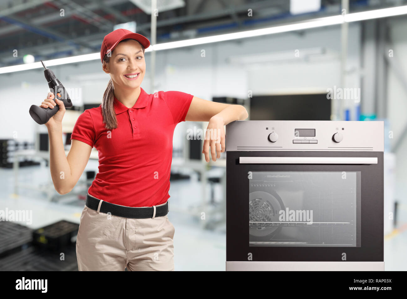 Repairwoman in una t-shirt rossa e un cappello rosso con un trapano e un forno in una fabbrica Foto Stock