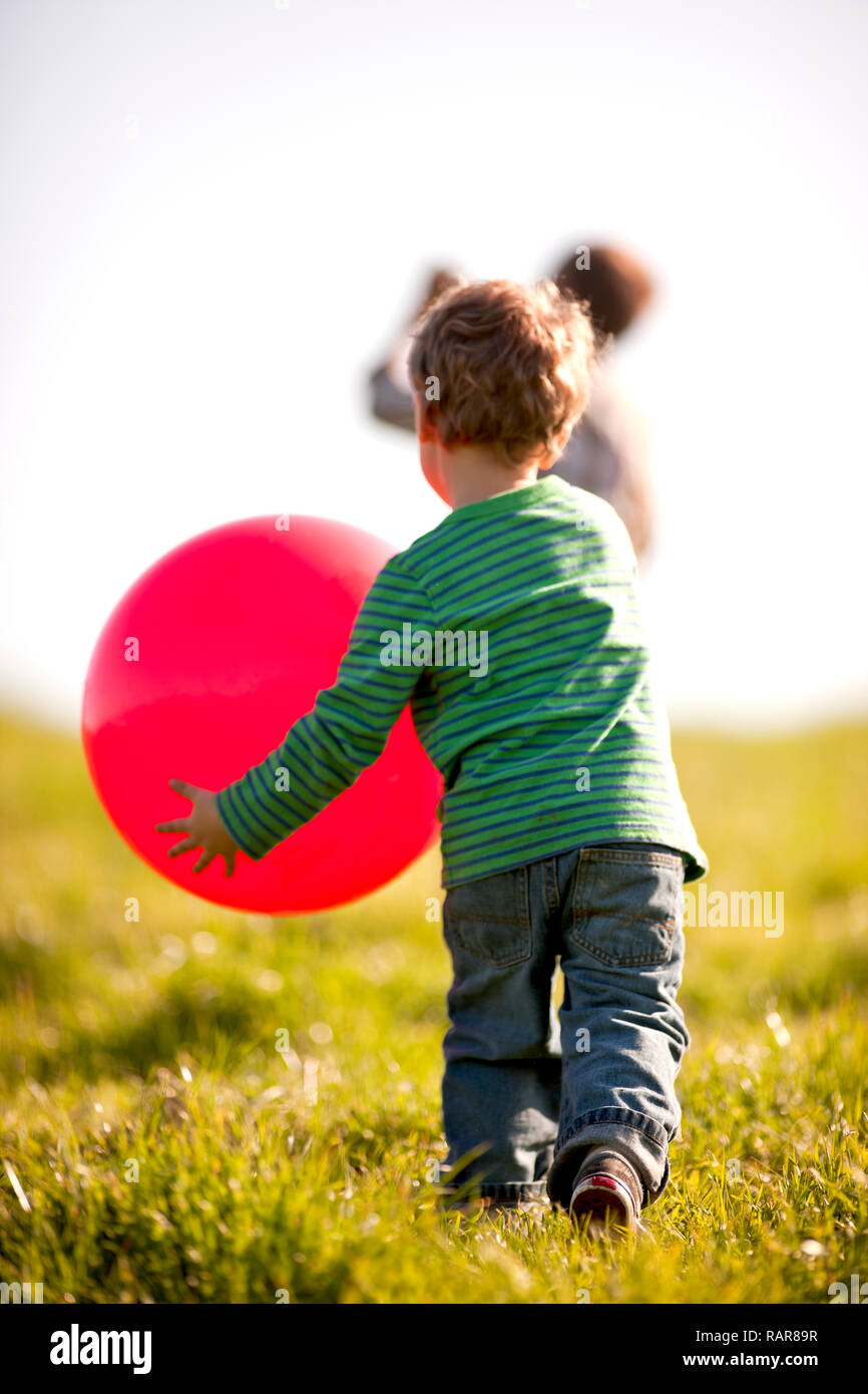 Vista posteriore di un ragazzo in esecuzione in un campo tenendo una grande sfera rossa. Foto Stock