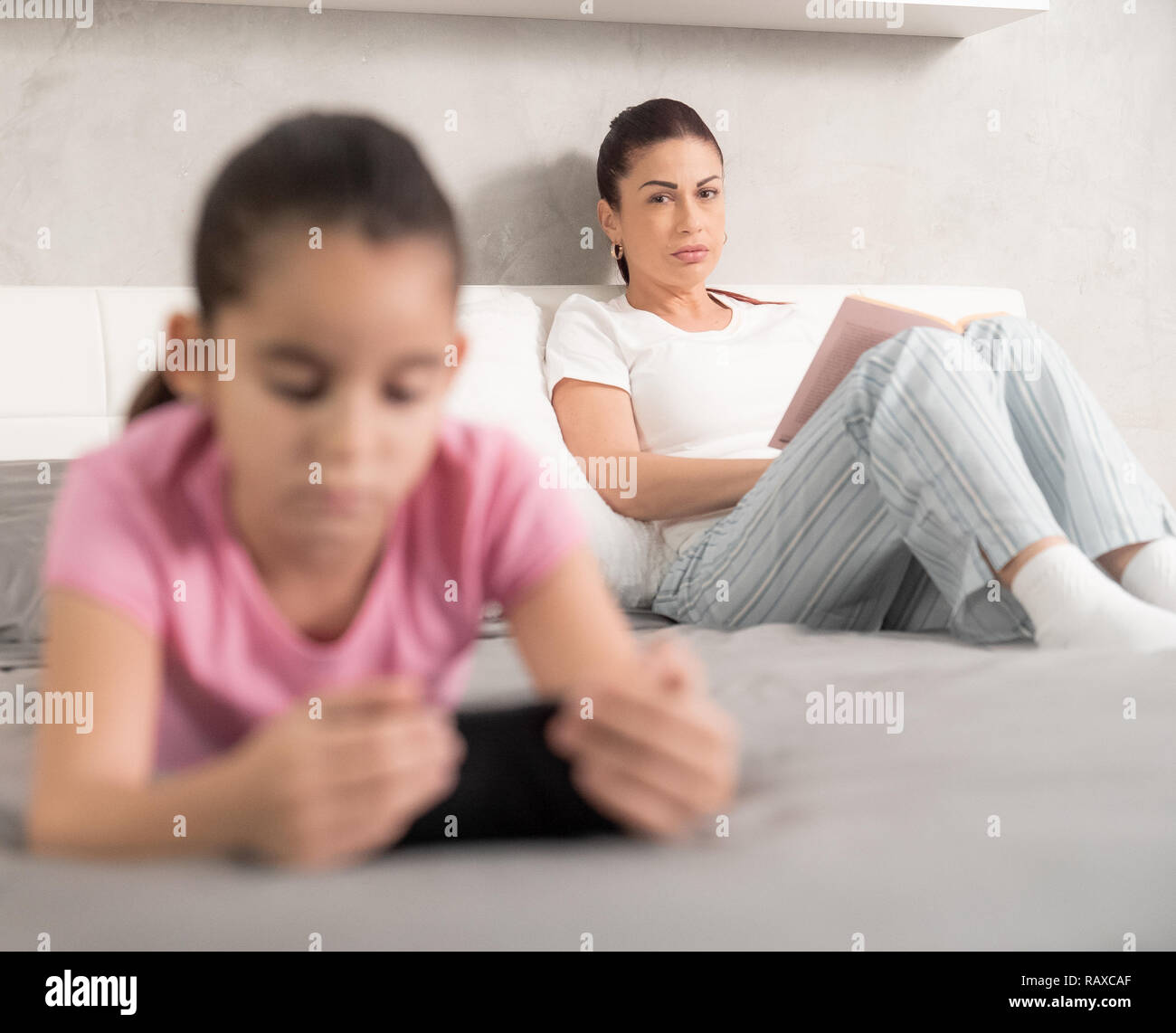 Ritratto di una singola bianco madre leggendo un libro guardando la figlia utilizzando il suo telefono mobile e di spendere troppo tempo su internet. Controllo principale della tecnologia e social media pericoli. ADHD Foto Stock