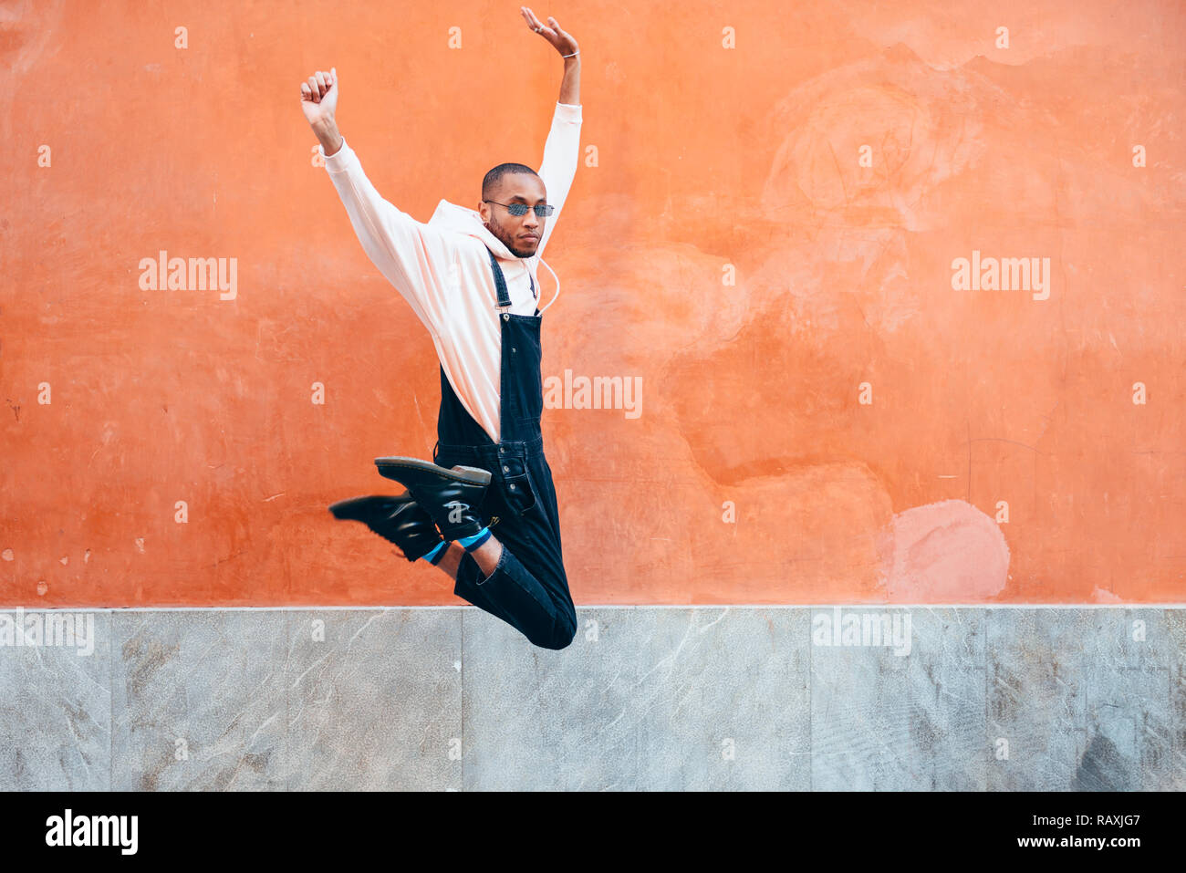 Giovane uomo nero indossando abiti casual jumping in background urbano. Il concetto di stile di vita. Millenaria ragazzo africano con bib pants all'aperto Foto Stock