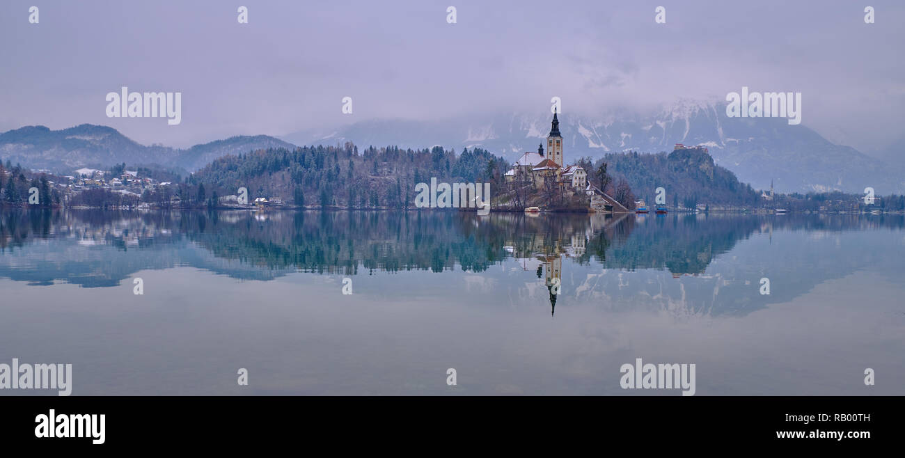 Inverno moody panorama lago di Bled shot, con la riflessione della Chiesa di pellegrinaggio dell Assunzione di Maria e la foresta e la montagna dietro Foto Stock