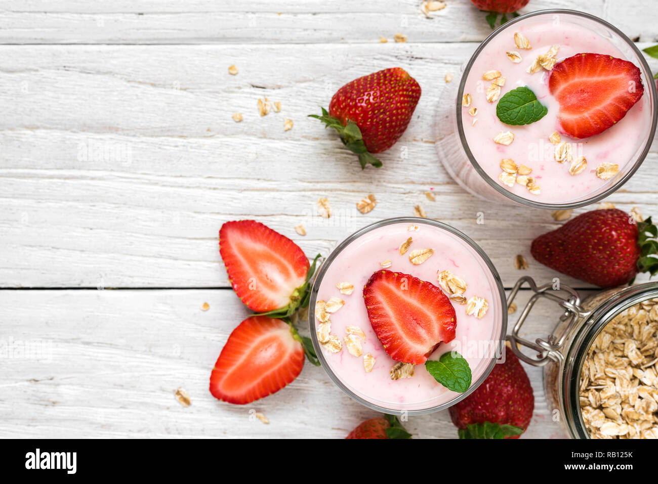 Due bicchieri di sano di yogurt alla fragola con frutti di bosco freschi, avena e menta su bianco tavolo in legno. Colazione sana. vista superiore con spazio di copia Foto Stock