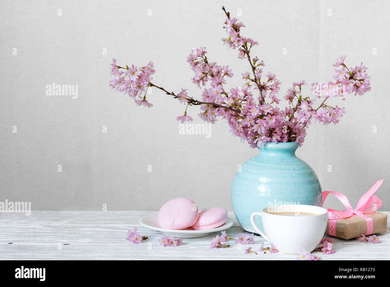 Rosa fiori di ciliegio bouquet di fiori con tazza di caffè, amaretti e confezione regalo in blu vintage vaso bianco su sfondo di legno. vacanze ancora vita Foto Stock