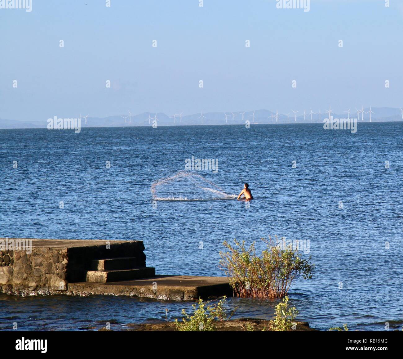 Un pescatore in piedi waist deep colata di una rete da pesca in lago di Nicaragua con grandi turbine eoliche in background Foto Stock