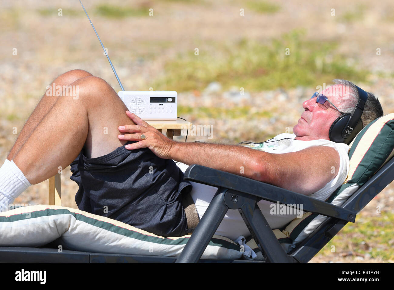 Uomo che stabilisce nel sole in una sdraio sulla spiaggia, indossando le cuffie per ascoltare la radio, durante l'estate 2018 ondata di caldo nel Regno Unito. Foto Stock