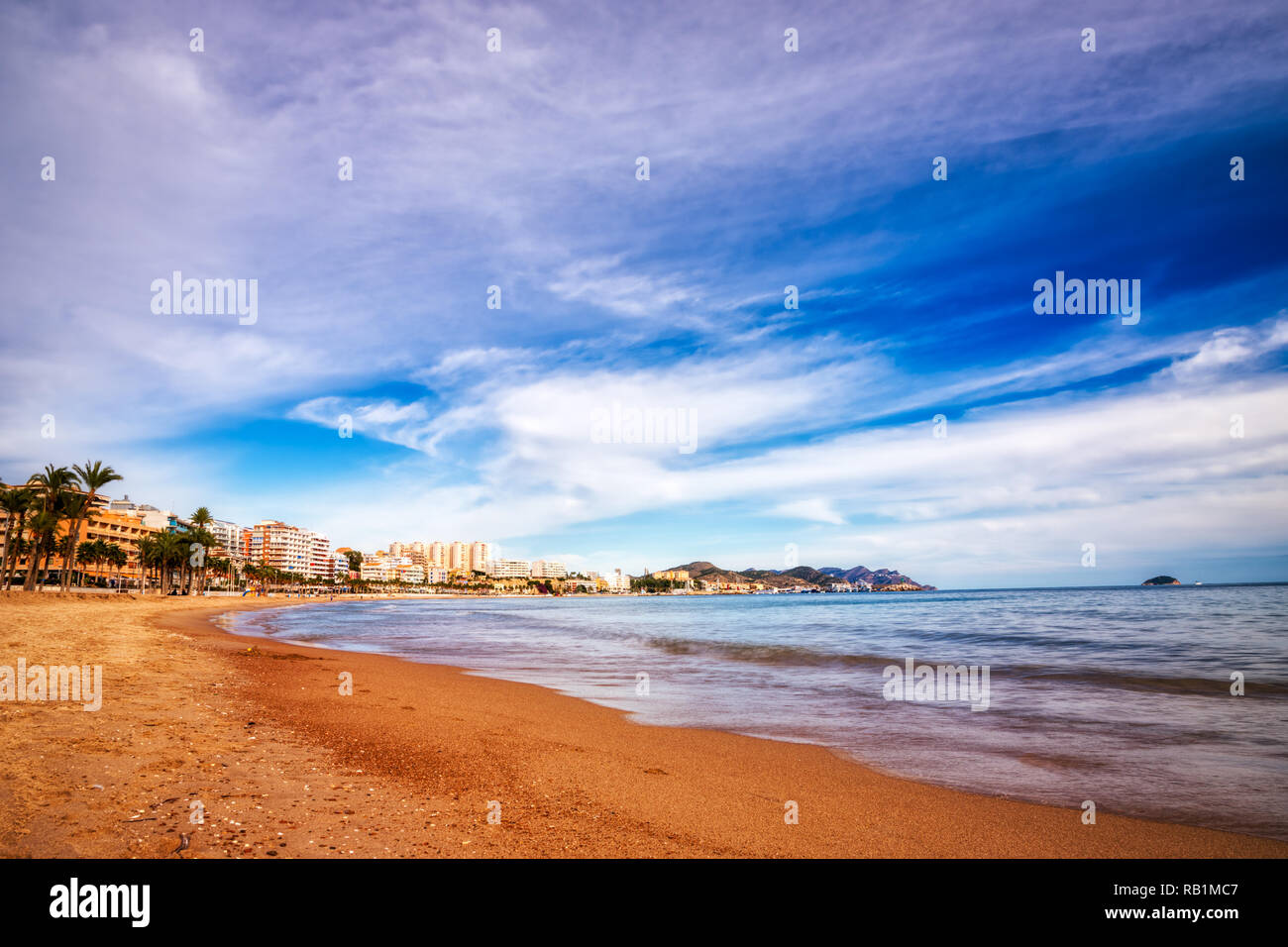 Bassa angolazione delle onde in arrivo sulla spiaggia snady di Villajoyosa spagna con un cielo blu, il bianco delle nuvole e gli edifici che fiancheggiano la riva in Foto Stock