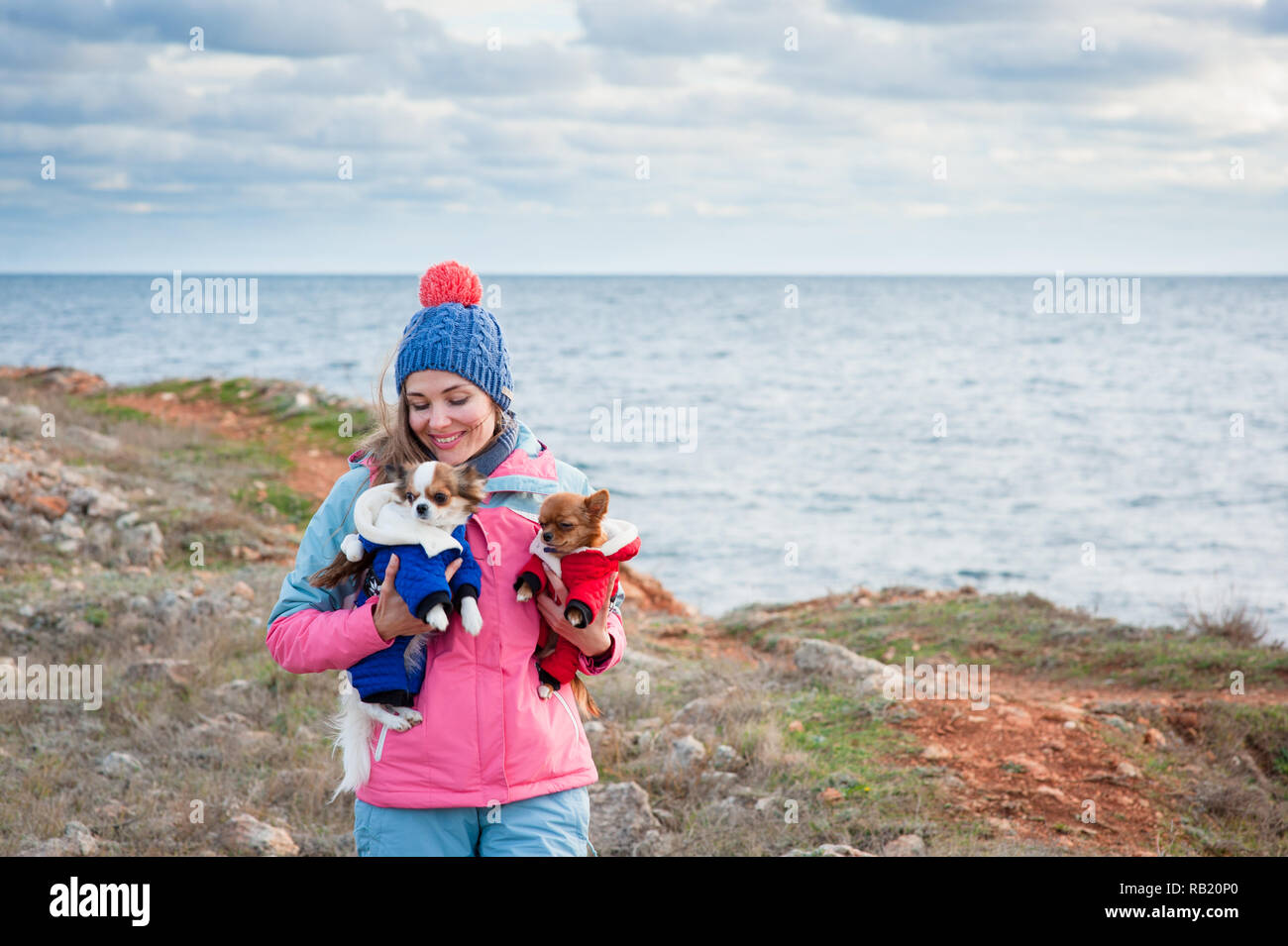 Felice delizioso sorridente piuttosto giovane femmina nel cappello di lana e tuta tenendo due piccole vestito in abiti animali chihuahua cani su freddo riva del mare in primavera Foto Stock
