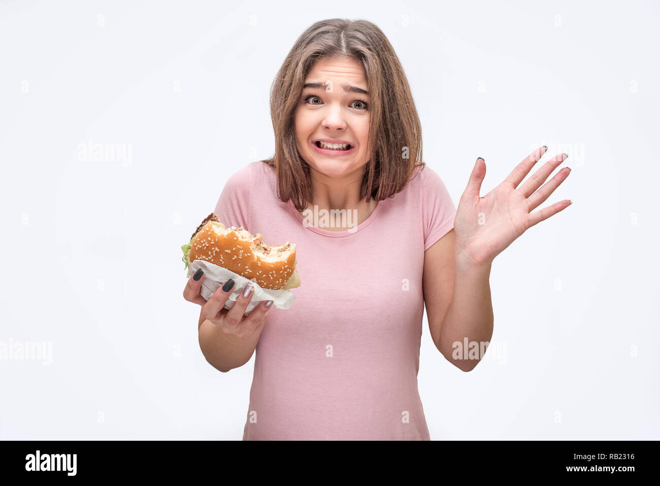 Spaventata e frustrati giovane donna attesa morso burger in mani. Ella mostra palm. Isolato su sfondo grigio. Foto Stock