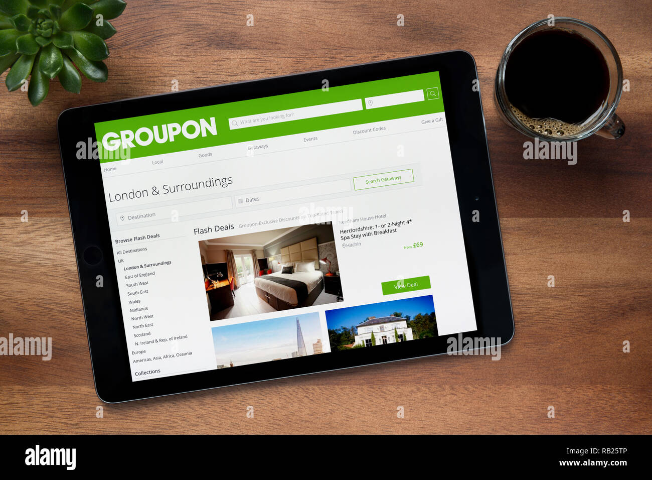 Il sito web di Groupon è visto su un tablet iPad, su di un tavolo di legno lungo con un caffè espresso e una pianta di casa (solo uso editoriale). Foto Stock