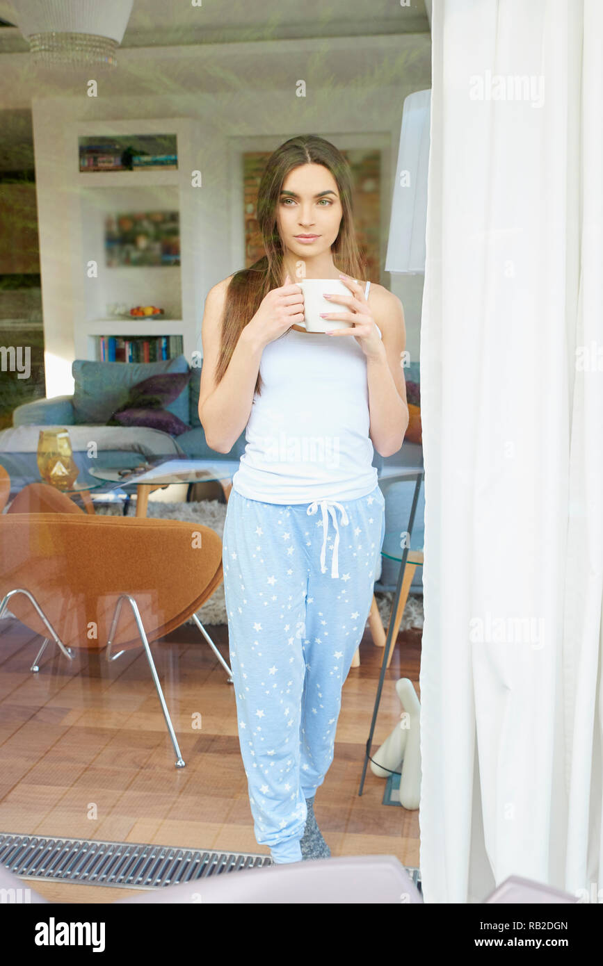 Ritratto di giovane donna graziosa indossando pigiami mentre in piedi con una tazza di caffè vicino alla finestra e sognare. Foto Stock