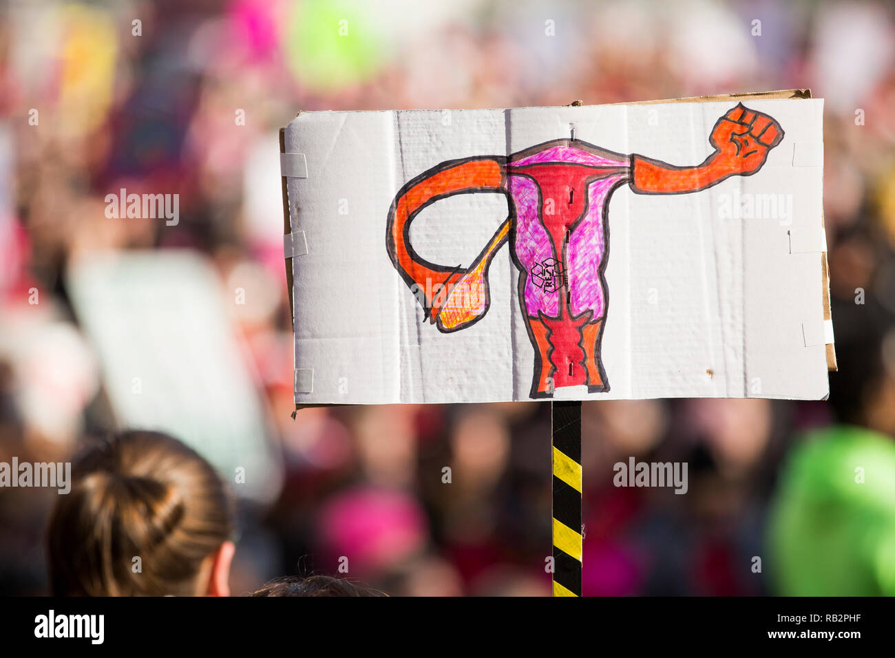 Disegnato a mano segno raffigurante donna organi riproduttivi formando un pugno di potenza sale oltre la folla durante il 2018 Donna marzo dimostrazione. Foto Stock