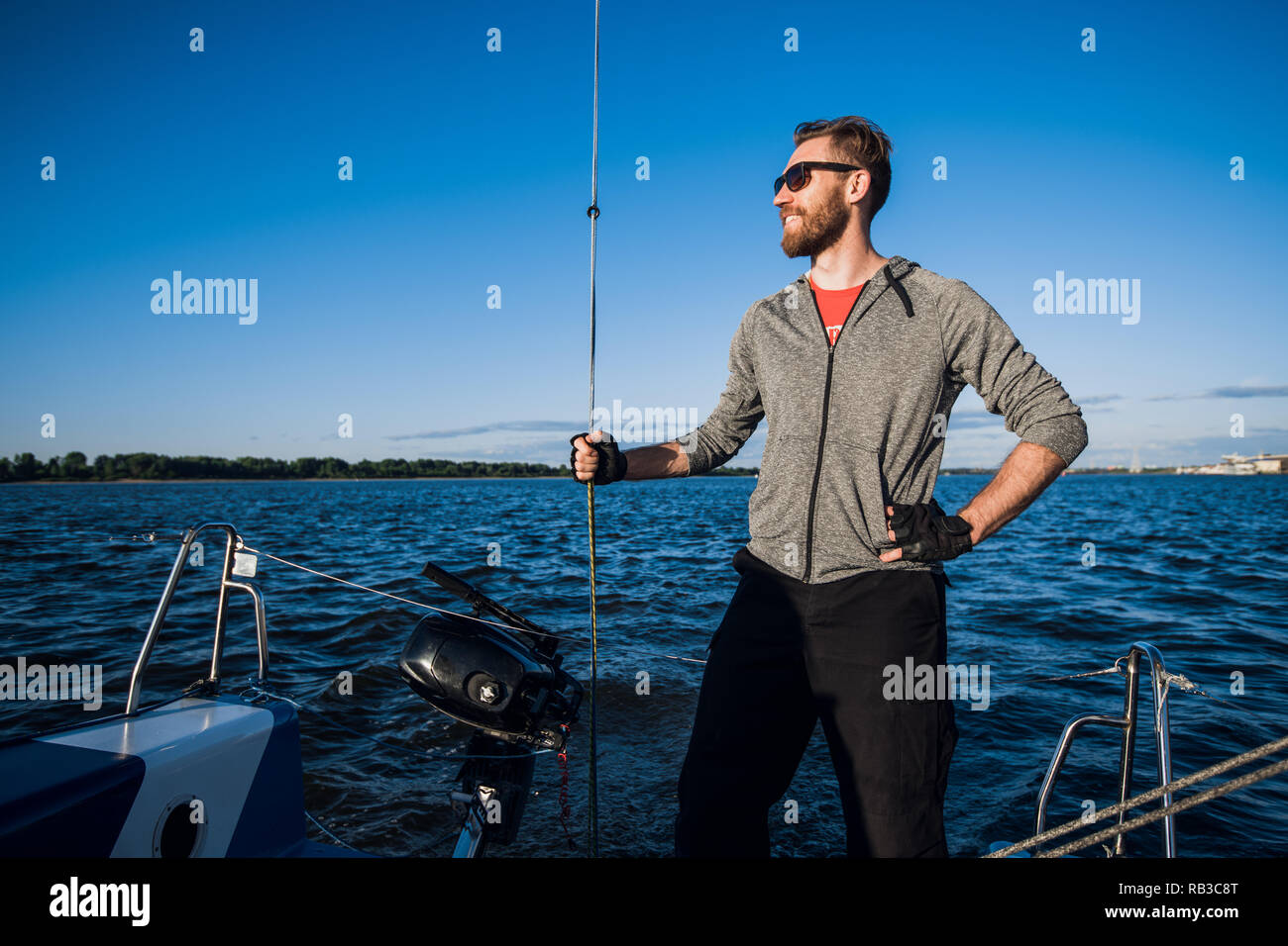 Giovane uomo che indossa gli occhiali da sole in piedi sulla poppa di yacht e godendo di una perfetta giornata autunnale sotto le vele - vacanze a vela nozione Foto Stock