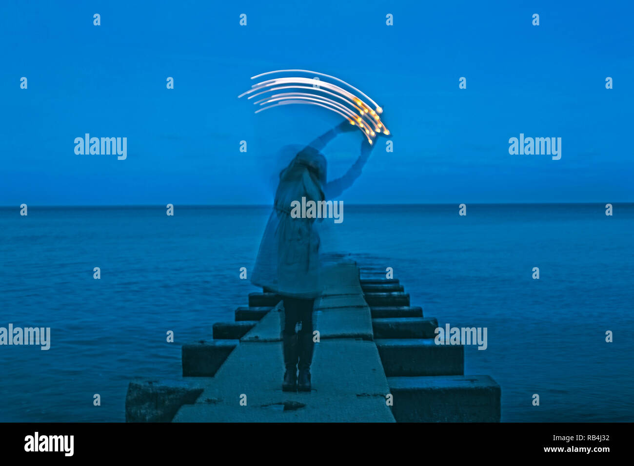 Una donna si erge su un molo tenendo una stella illuminata sopra la sua testa, spostandolo come una stella cadente, al crepuscolo Foto Stock