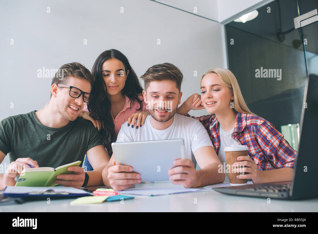 Bello e attraente ai giovani si siedono a tavola. Essi guardano a tavoletta e sorriso. Brunette stnd dietro di loro. Essi lavorano insieme. Foto Stock