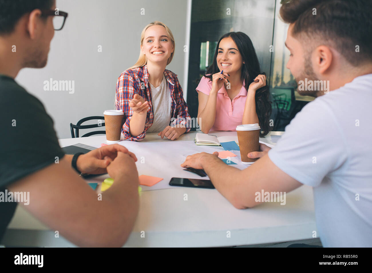 Felice e positiva delle giovani donne guardare l uomo e il sorriso. Essi hanno tazza di caffè al tavolo. Persone sedersi insieme in camera bianca. Essi lavorano. Foto Stock