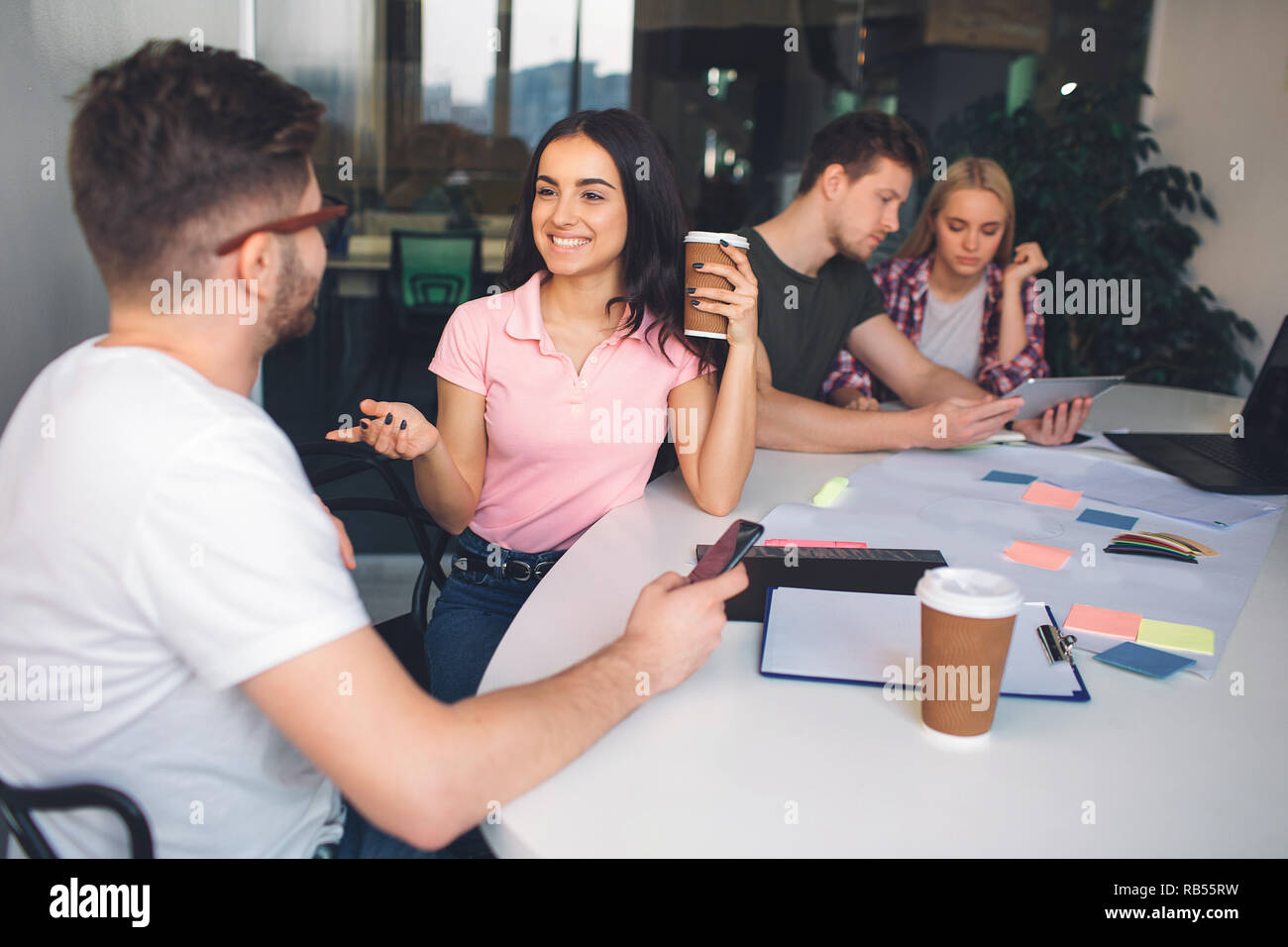 Felice e positivo bruna giovane guardare guy e sorriso. Ella tenere tazza di caffè. Egli guarda a lei. Un altro paio di sedersi dietro di loro e lavorare insieme. Foto Stock