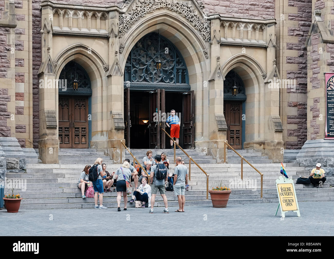 Gruppo di persone sedute davanti a San Giacomo Regno chiesa protestante dietro la Chiesa segno aperto con un lgbt bandiera arcobaleno Foto Stock