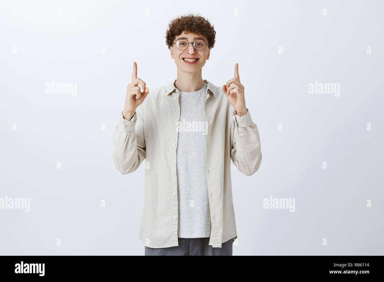 Affascinante ottimista e emotivo giovane bello giovane maschio in bicchieri arrotondati e capelli ricci alzando le mani per puntare in alto e sorridente orgogliosamente indossando occhiali da stupidi e camicia oltre il muro grigio Foto Stock