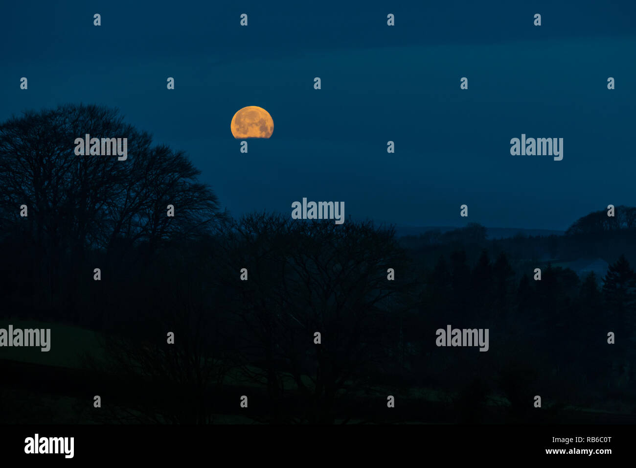 Uno scenario naturale di una luna piena che salgono attraverso una pausa nelle nuvole sopra un oscuro paesaggio rurale con alberi Foto Stock
