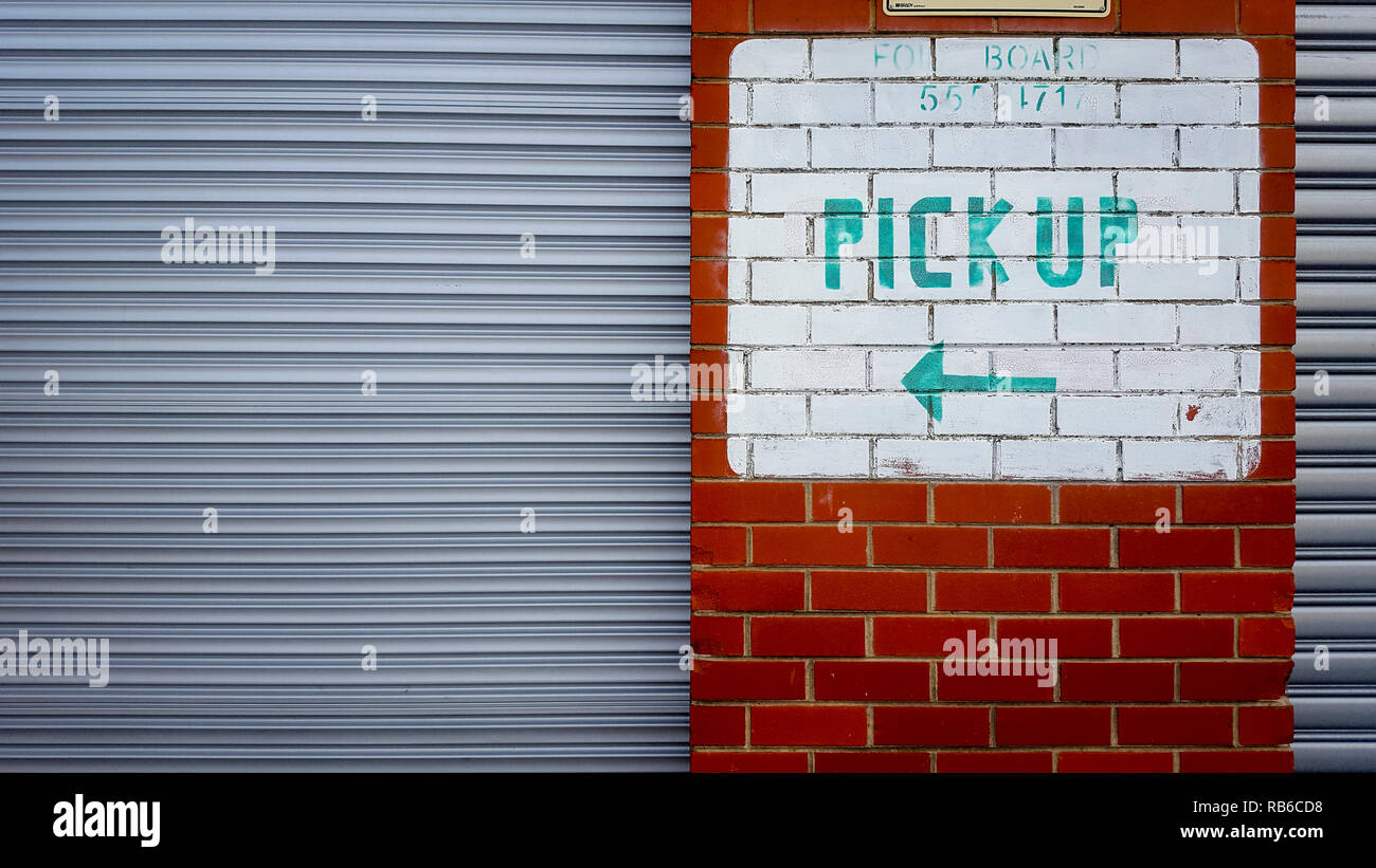 Un vecchio sbiadito Pick Up segno con una freccia, il testo in verde contro pitturato di bianco su rosso di un muro di mattoni con argento porte a rullo Foto Stock