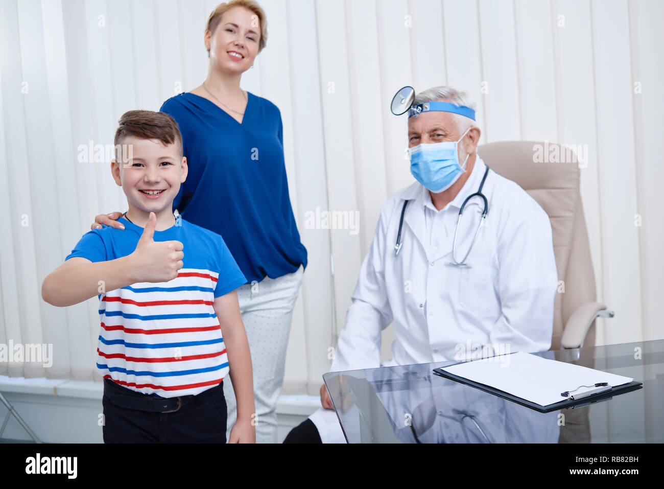 Ritratto di albero la gente in ospedale. La mamma con suo figlio in piedi sorridente a guardare fotocamera dopo consultazione professionale in ospedale. Medico seduto dietro il tavolo in maschera e uniforme nel suo ufficio. Foto Stock