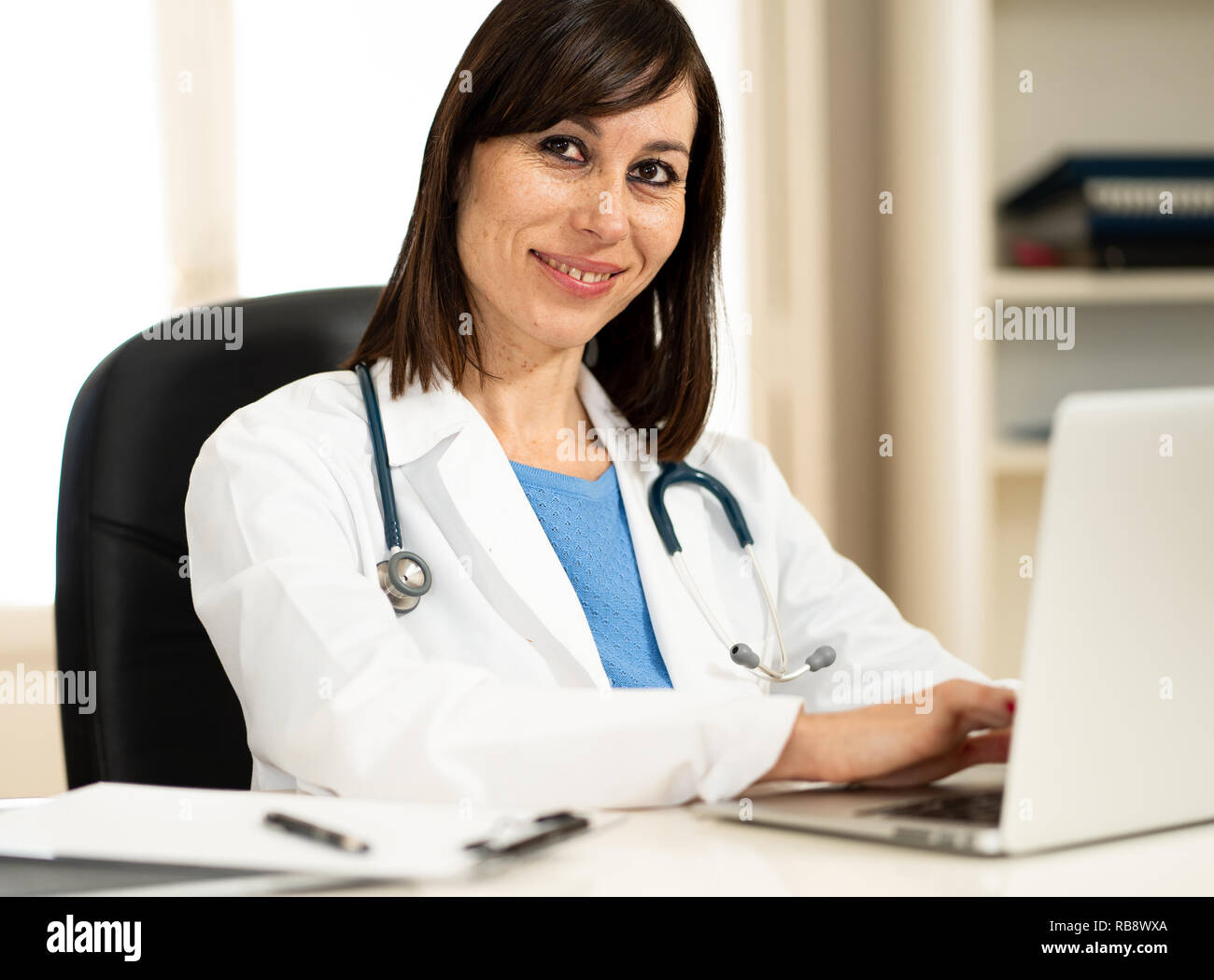 Ritratto di felice donna attraente medico in camice bianco a lavorare con il computer portatile e la scrittura sulla documentazione seduta su clinic hospital scrivania in ufficio Foto Stock