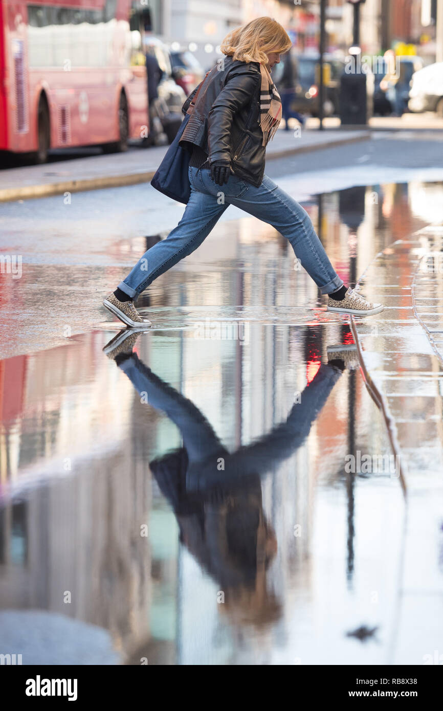 Una donna passi al di sopra di una pozza d'acqua stagnante su Oxford Street, Londra, a seguito di una raffica acqua principali in uno della capitale più trafficate strade dello shopping. Foto Stock