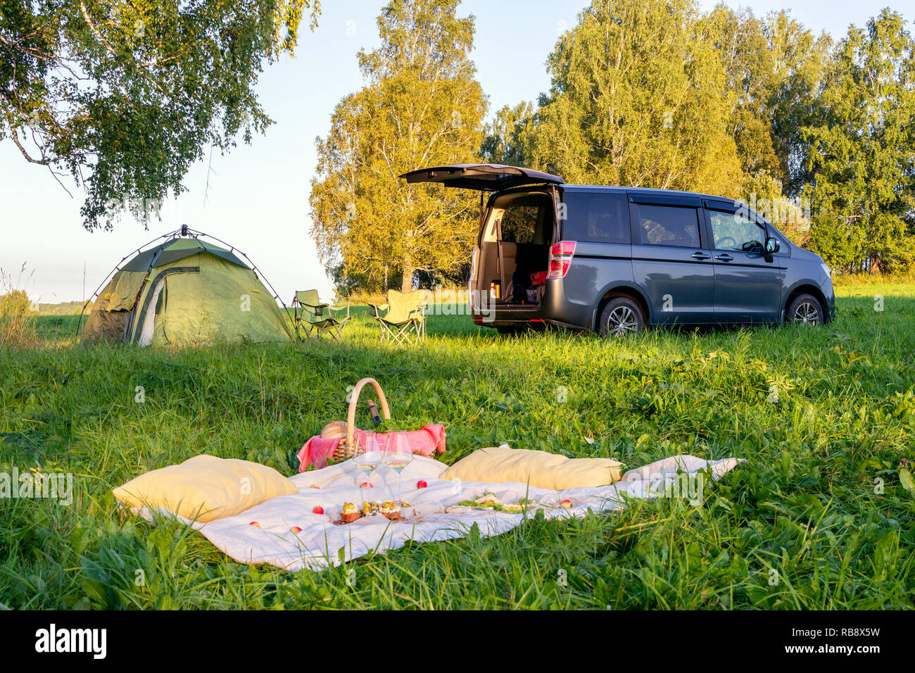 Picnic in foresta, tenda e camp sedie, grigio minibus con porta aperta, coperta, cesto di vimini, bicchieri da vino, snack, frutta, pane, cuscini. Concetto Foto Stock