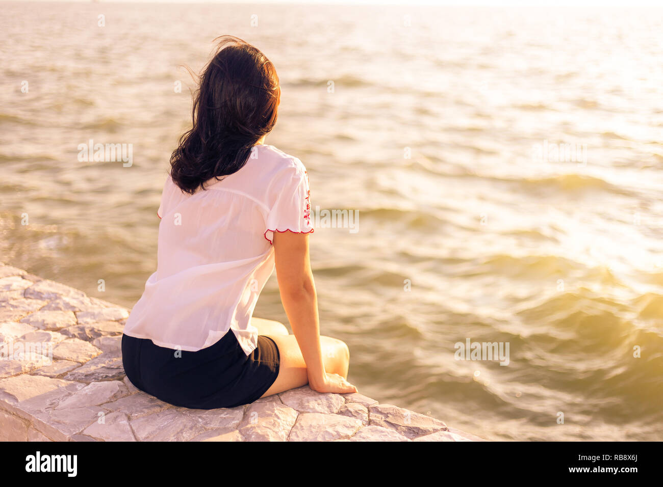 Giovane donna seduta sul muro di pietra che si gode a guardare il tramonto sulla spiaggia, ottimo per rilassarsi o rallentare il concetto di vita Foto Stock