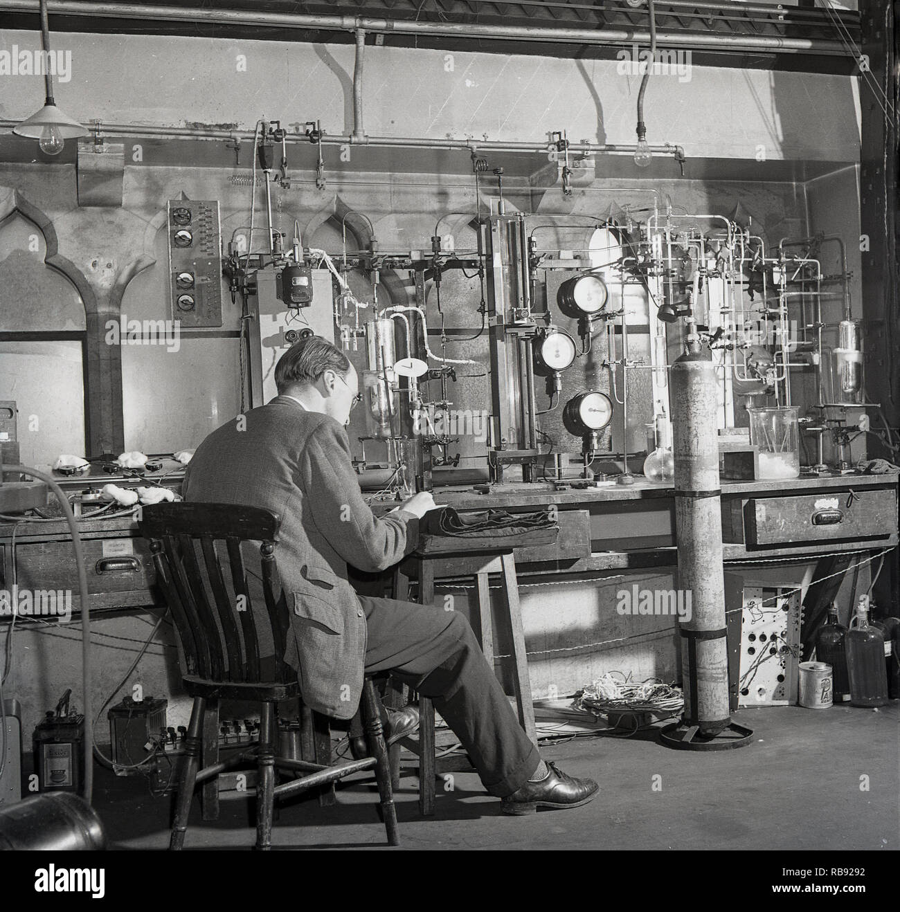 Anni '1950, storico, professore maschio seduto in un laboratorio scientifico presso l'Università di Oxford, Inghilterra, Regno Unito. Sta conducendo esperimenti utilizzando un'ampia varietà di attrezzature, tra cui batterie, tubi di gomma, taniche di gas, tubi e manometri. Foto Stock
