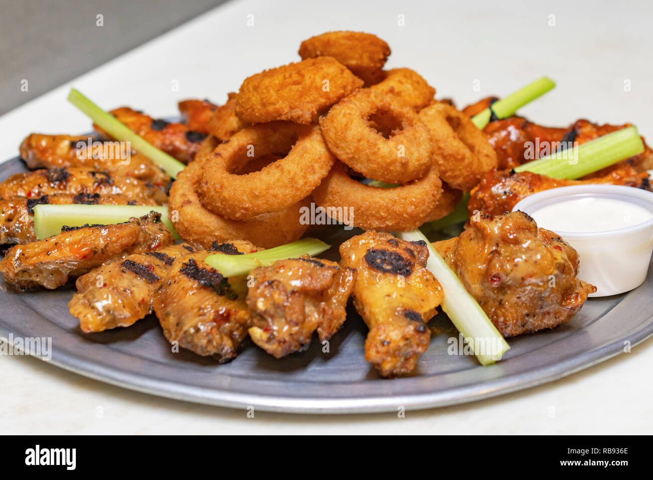 Il cibo da bar: Ala di pollo piatto con anelli di cipolla e sedano. Contenitore di bianco salsa di immersione sul pannello bianco. Vista laterale vicino. Foto Stock