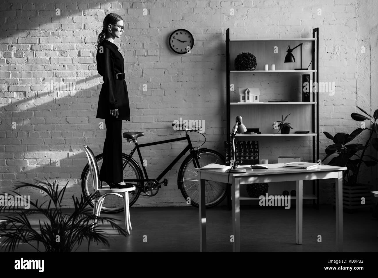 In bianco e nero di donna attraente vestiti di nero permanente sulla sedia bianca vicino a bike Foto Stock