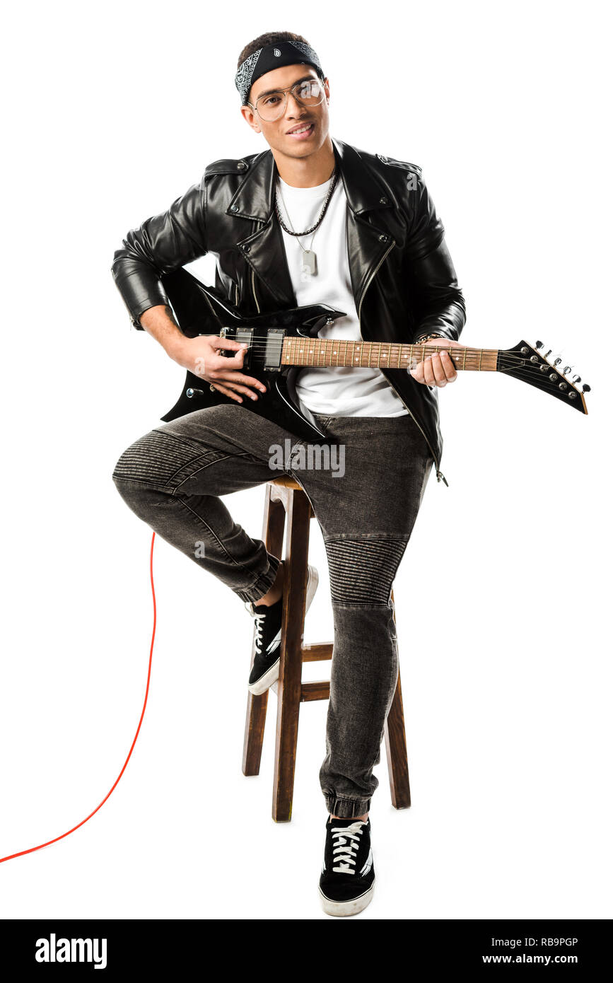 Allegro maschio musicista rock in giacca di pelle giocando sulla chitarra elettrica mentre è seduto su una sedia isolato su bianco Foto Stock