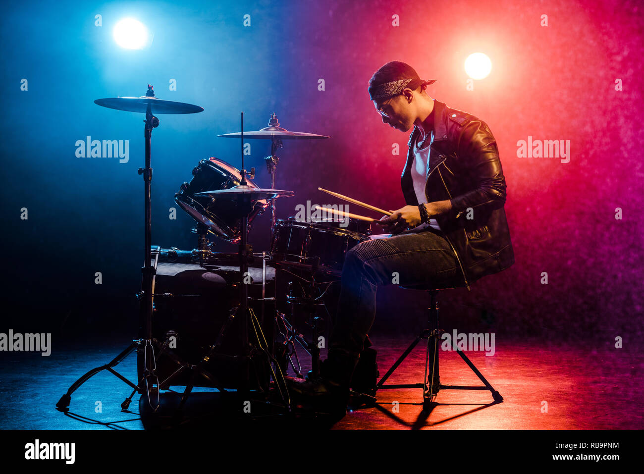 Vista laterale del musicista maschio in giacca di pelle suonando la batteria durante un concerto rock sul palco con fumo e faretti Foto Stock