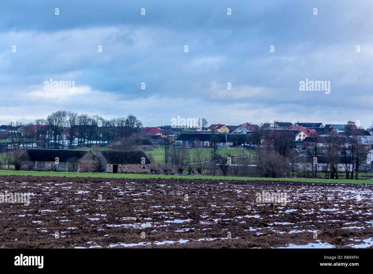 Un po' di neve sul campo arato. Villaggio e alberi in background. L'inizio dell'inverno in Europa. Foto Stock