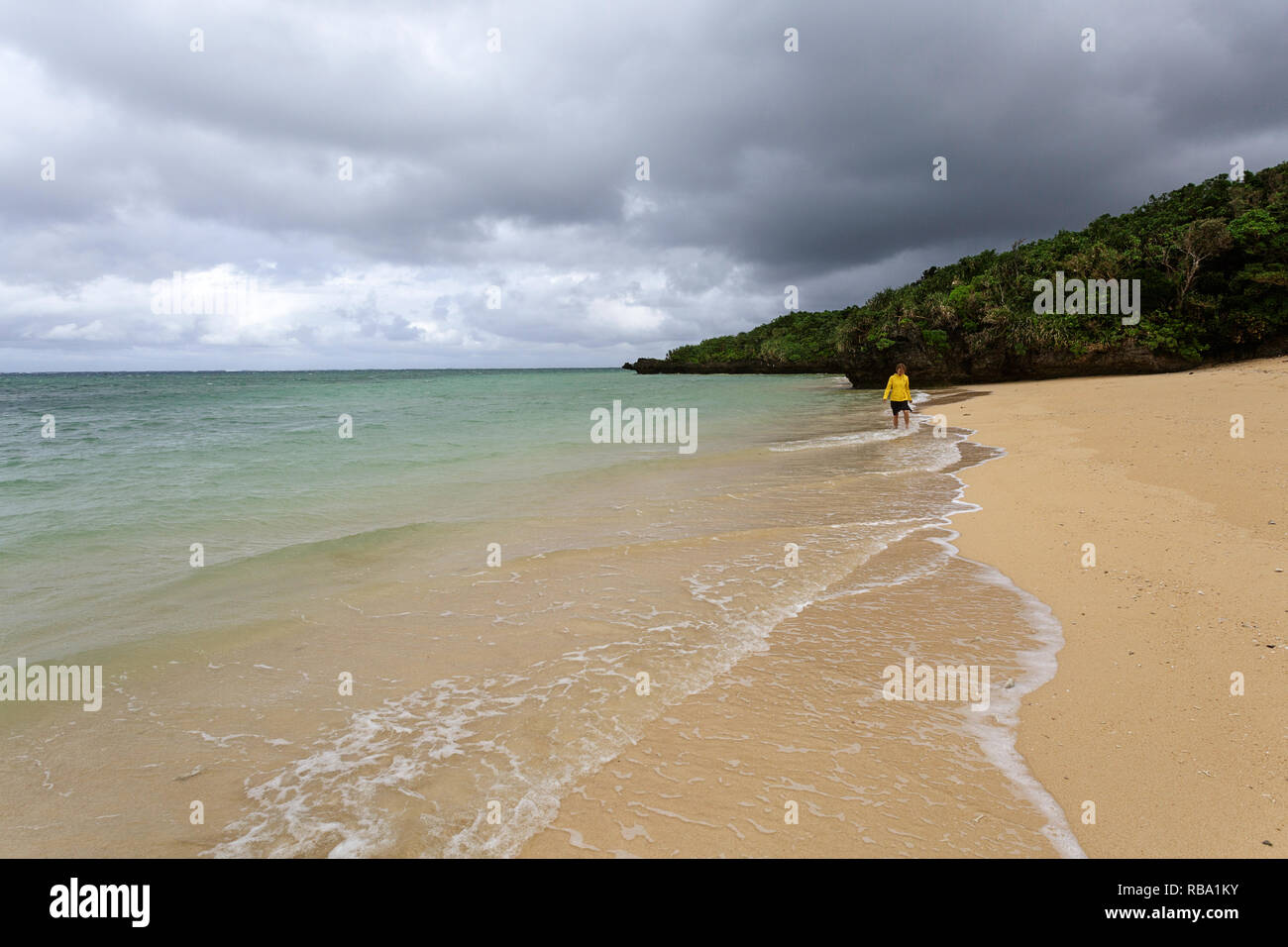 Giovane donna in giacca gialla in piedi, camminare in acque poco profonde su una bellissima spiaggia appartata, Ishigaki, Giappone Foto Stock