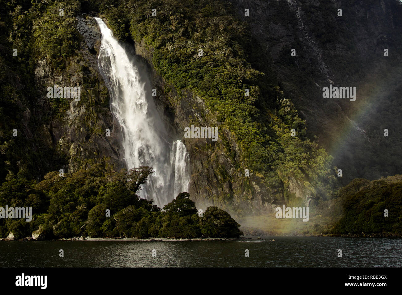 Milford Sound con cascata arcobaleno dalla base. Sun mette in evidenza la cascata, rocce e cliff Foto Stock