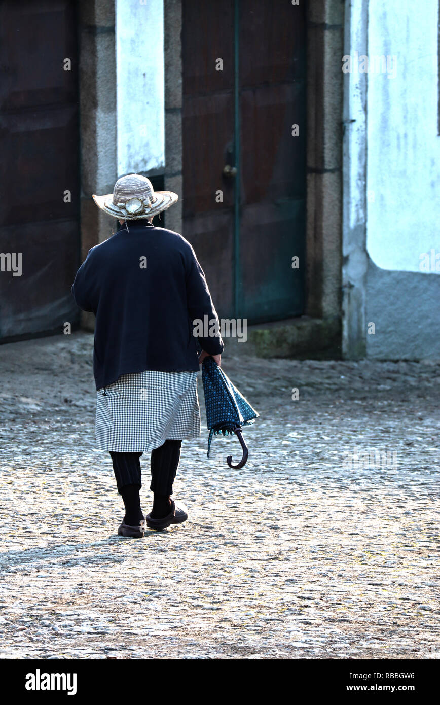 Un pomeriggio invernale in Vila Cova de Alva. Una signora anziana cammina giù per la collina dalla chiesa. Il umberella ombreggiatura era lei da sole d'inverno. Foto Stock