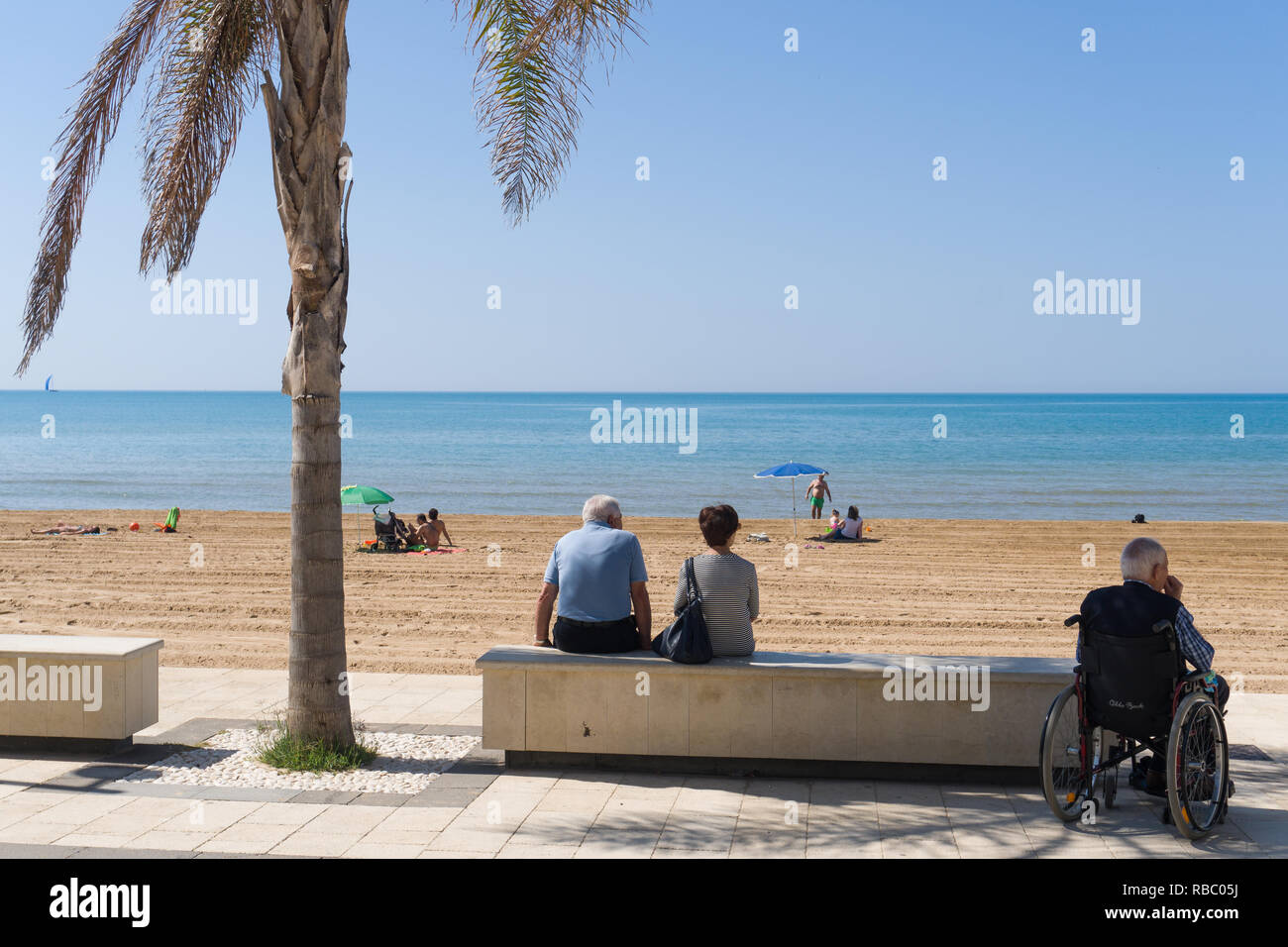 Coppia senior e sedie a rotelle utente seduto in ombra e guardare le famiglie sulla spiaggia Foto Stock