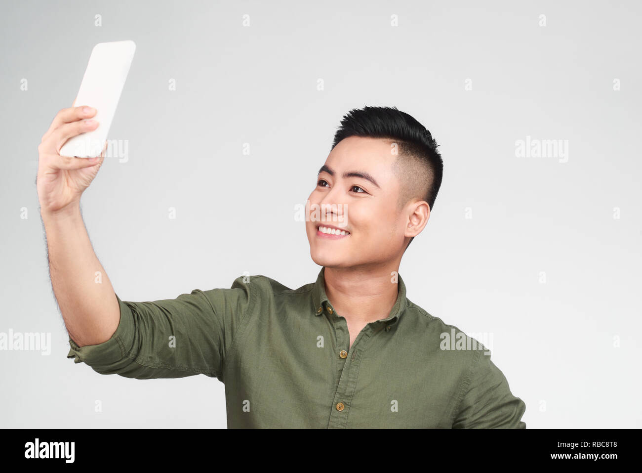 Bel ragazzo in camicia bianca si sta facendo selfie utilizzando uno smart phone, mostrando Ok segno e sorridente, su sfondo grigio Foto Stock