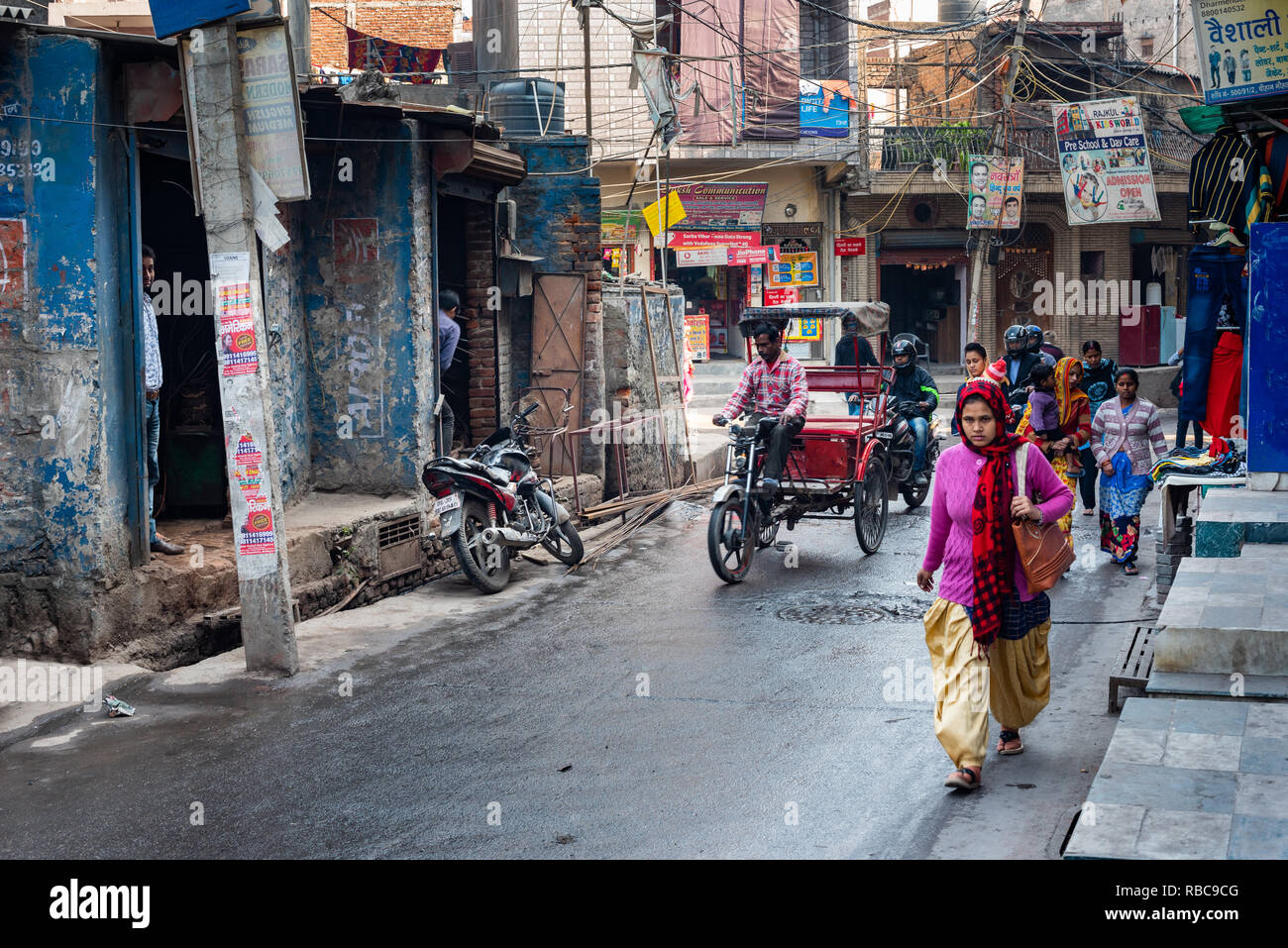 Una strada trafficata in Madanpur Khadar villaggio con e-rickshaws, due ruote e pedoni su una strada bagnata fiancheggiata da negozi con un drain aperto. Nuova Delhi Foto Stock
