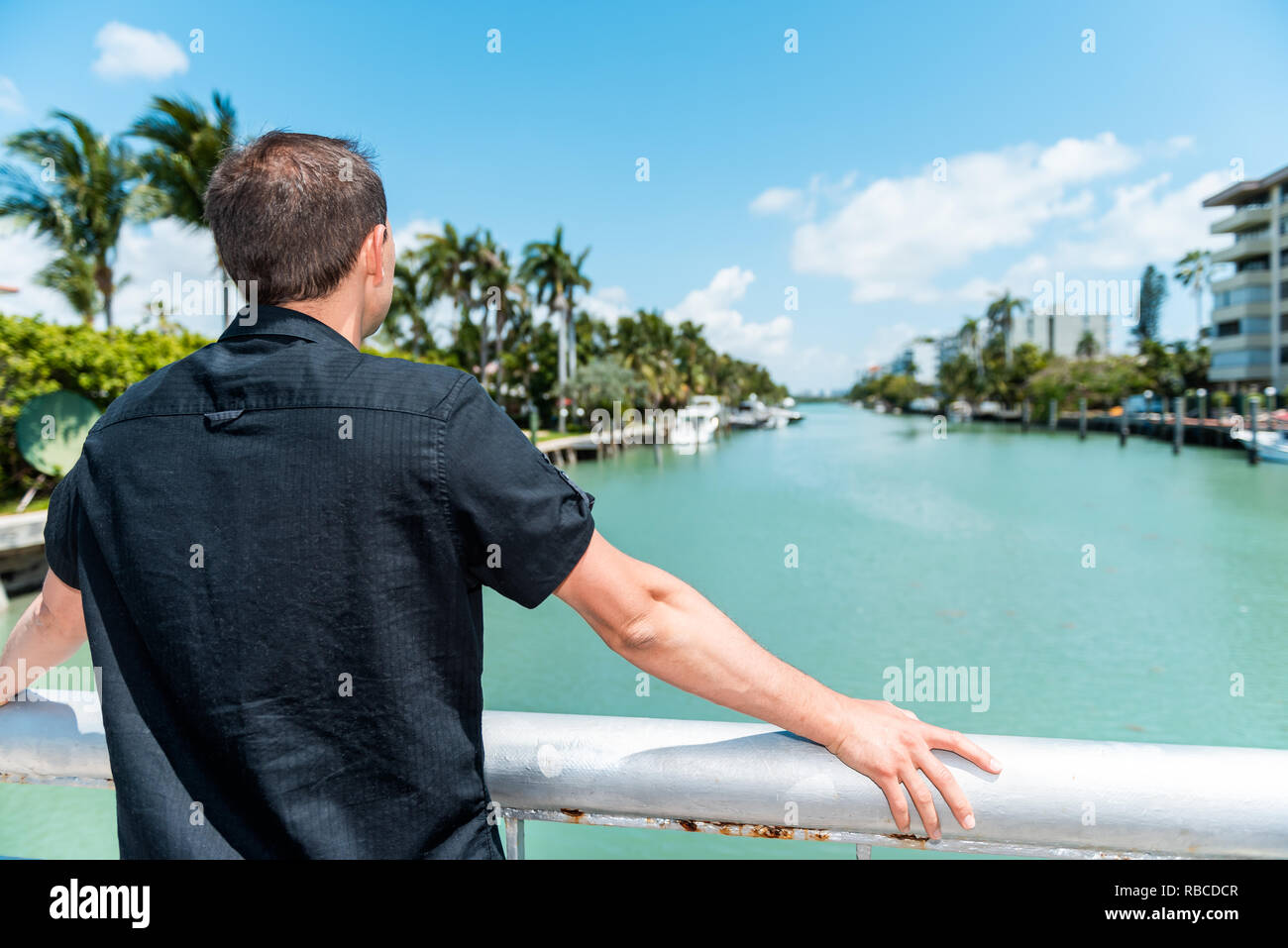 Giovane uomo torna indossando la maglia nera in piedi appoggiata sulla ringhiera del ponte in Bal Harbour, Miami Florida con oceano verde della Baia di Biscayne Foto Stock