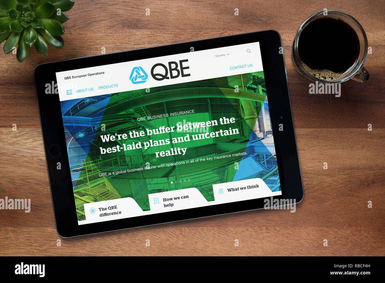 Il sito web di QBE Insurance Group è visto su un tablet iPad, su di un tavolo di legno lungo con un caffè espresso e una pianta di casa (solo uso editoriale). Foto Stock