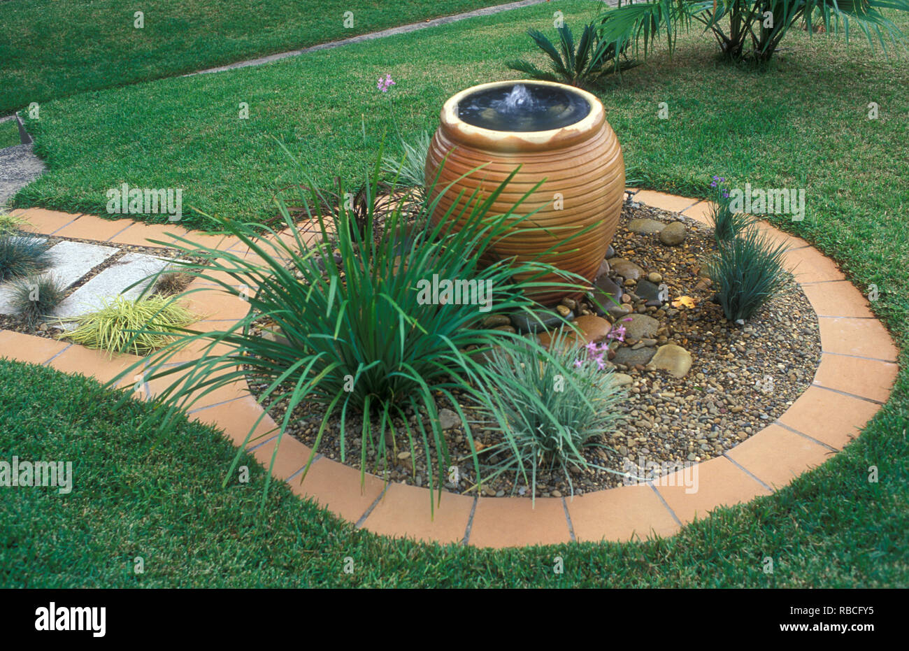Circolare giardino di ghiaia, mattonelle in terracotta, bordatura contenitore fontana, SISYRINCHIUM, TULBAGHIA VIOLACEA, LOMANDRA E FESTUCA BLU (Festuca glauca) Foto Stock