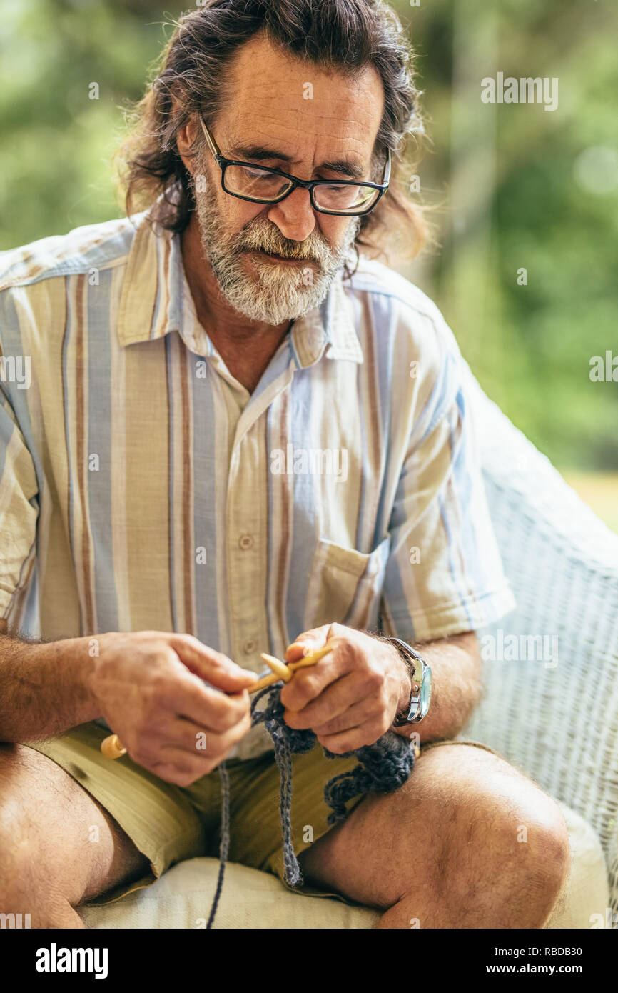L'uomo anziano con la barba maglia mentre è seduto sulla sedia. Il vecchio uomo maglia con aghi e filati di lana. Foto Stock