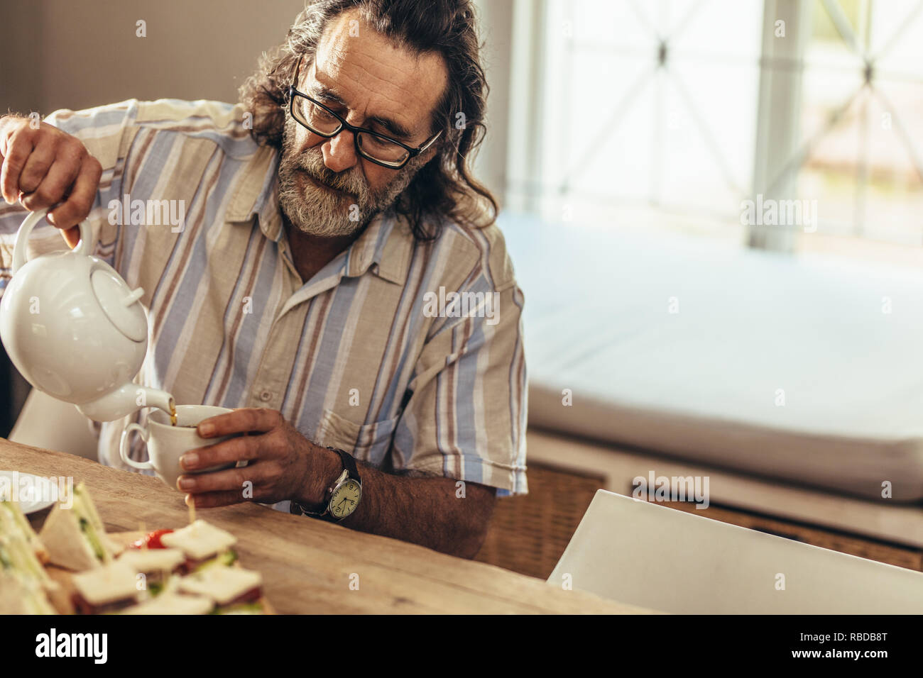 Uomo anziano con la barba versando il tè in dalla teiera alla tazza seduti a tavola all'interno. Uomo in pensione che serve una tazza di tè. Foto Stock