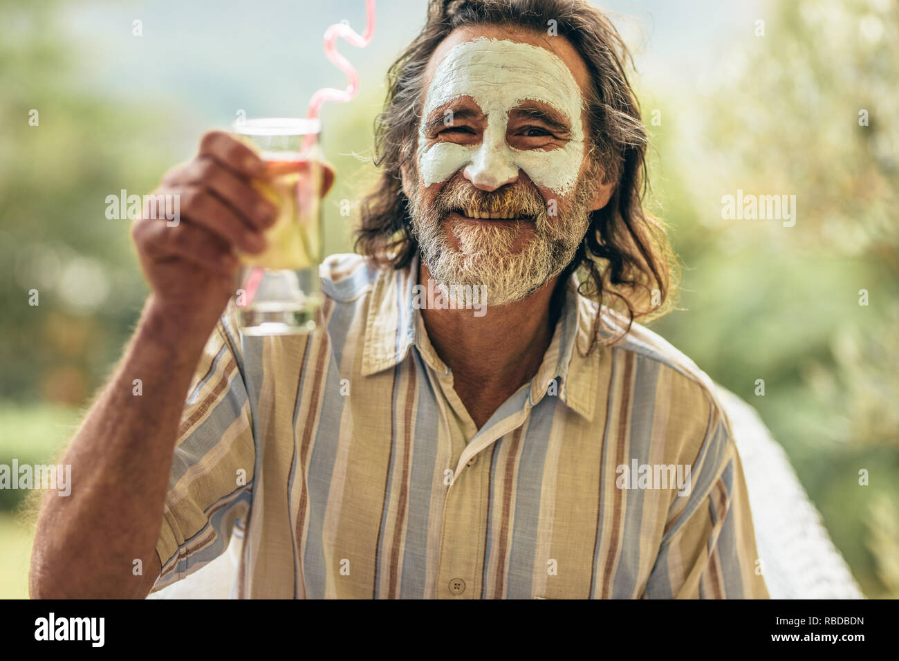 Senior barbuto uomo con argilla maschera facciale tenendo un bicchiere di succo di frutta in mano. Man aventi spa trattamento viso sollevando il suo succo vetro alla fotocamera. Foto Stock